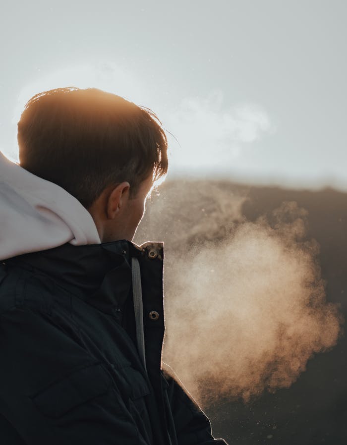 A man in a jacket exhales visible breath in the cold outdoor sunlight, creating a serene winter scene.